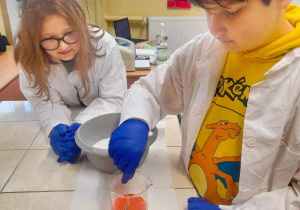 Na zdjęciu chłopiec i dziewczynka ubrani w fartuchy laboratoryjne i niebieskie rękawice, znajdujący się przy stole laboratoryjnym. Na pierwszym planie chłopiec mieszający pomarańczowy płyn w zlewce, z tyłu asystująca przy doświadczeniu uczennica.