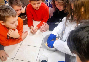 Na zdjęciu grupa dzieci stojąca dookoła stołu laboratoryjnego , dwoje z nich, ubranych w fartuchy i niebieskie gumowe rękawice miesza w plastikowej misce masę, dodając do niej kolejne barwniki. Pozostała część dzieci obserwuje doświadczenie.