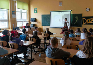 Zdjęcie przedstawia salę, a w niej klasa czwarta, która bierze udział w warsztatach o kulturze Słowian. Uczniowie przysłuchują się opowieści, które snuje stojąca przed nimi uczennica XXI Liceum - Maria, ubrana w tradycyjny, ludowy strój opoczyński.