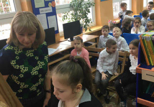 Zdjęcie na pierwszym planie ukazuje panią bibliotekarkę razem z uczennicą, która zapisuje pod okiem pani tekst na tablicy. W tle siedząca na krzesełkach, ubrana na galowo grupa dzieci.
