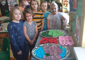 Zdjęcie przedstawia grupę dzieci stojących obok stolika, na którym znajdują się wcześniej wykonane przez nie pachnące, kolorowe mydełka.