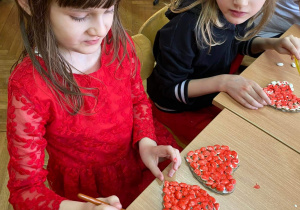 Zdjęcie przedstawia dwie dziewczynki, które malują przyklejone na kartonowym sercu fasolki czerwoną farbą.