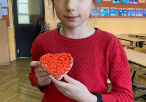 Na zdjęciu dziewczynka trzymająca w rękach wykonane z fasolek czerwone serduszko walentynkowe.