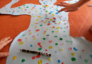Zdjęcie przedstawia wycięty z brystolu kształt człowieka z podniesionymi rękami, a dookoła znajdują się dzieci i palcami umoczonymi w kolorowych farbach stęplują wycięty brystol.