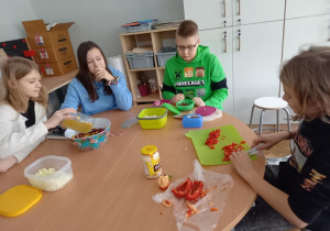 Grupa uczniów siedzi przy okrągłym stoliku i przygotowuje sałatkę. Na ławce leżą składniki do potrawy oraz akcesoria kuchenne.