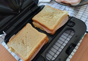 Zdjęcie czarnego opiekacza, w którym są przygotowane tosty z chleba. W oddali widać talerz z wędliną.