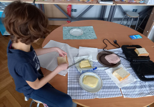 Dziewczynka siedzi na stołku i smaruje chleb masłem. Na ławce przygotowane składniki do robienia tostów oraz czarny opiekacz.