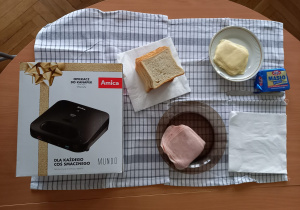 Zdjęcie składników do robienia tostów oraz opiekacza, zapakowanego w pudełko z napisem Amica. Produkty leżą na ściereczce w niebieską kratkę.