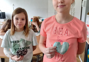 Dwie dziewczynki prezentują wypchane serca z tkaniny. W oddali widać salę lekcyjną.