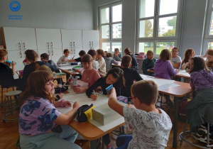 Zdjęcie grupy uczniów, siedzących w ławkach i malujących pudełka kartonowe. W oddali widać duże okna sali lekcyjnej.
